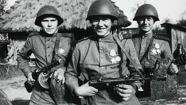 Three proud young soldiers posing in front of thatched hut